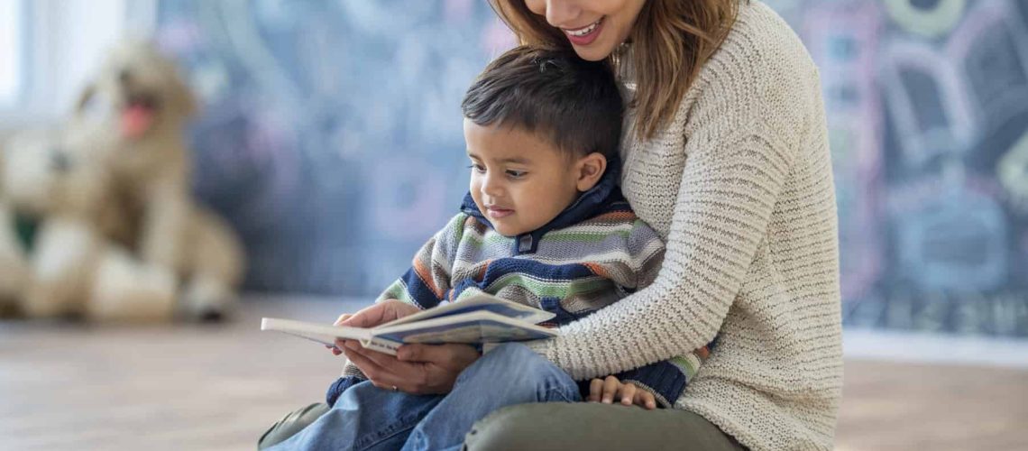 An Indian mother and son are seated together on the floor of their living room. The mother is about to read a story to his son who is sitting on her lap.